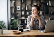 Tasse de café fumante posée sur un bureau en bois clair, avec un ordinateur portable ouvert et des documents légèrement éparpillés, suggérant le stress. La lumière est douce et naturelle, créant une ambiance studieuse.